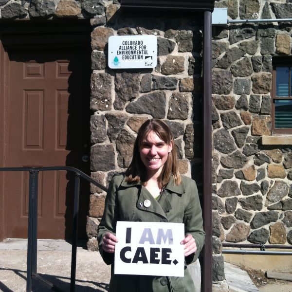 Katie smiles and holds a sign that reads "I AM CAAEE." in front of a stone building. 